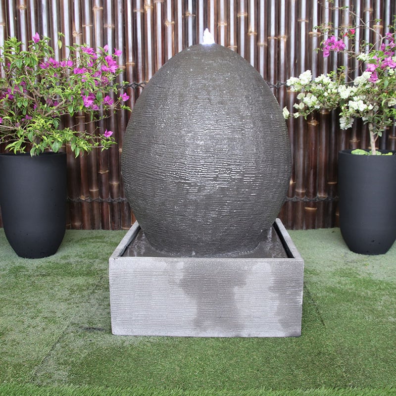 Galleria Imports Egg Fountain in grey front view with flowers either side